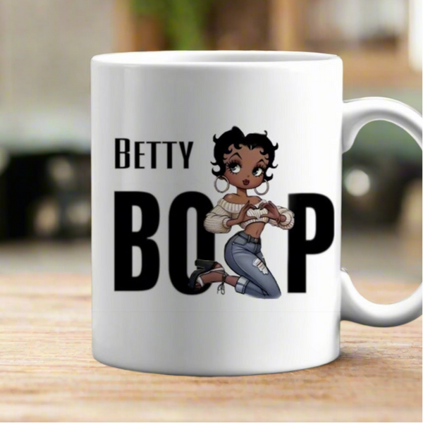The Betty Boop Ceramic Mug by Kreative Kreationz is a white 11oz ceramic mug with "Betty Boop" text and a cartoon of Betty Boop kneeling, set on a blurred indoor background. Dishwasher safe.