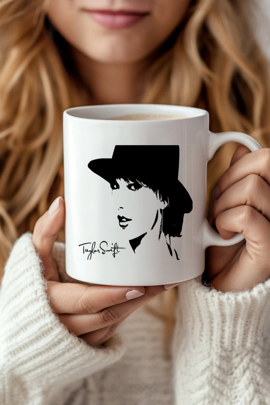 A person with long blonde hair, wearing a cozy white sweater, holds a Kreative Kreationz Taylor Swift Mug. The ceramic mug features a stylized illustration of a woman in a hat and signature-like text below the design.
