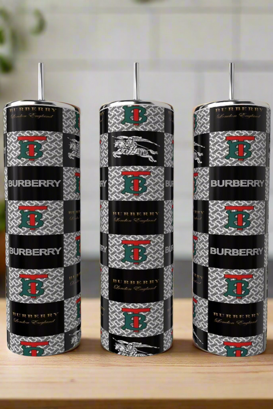 Three Burberry Inspired 20oz Tumblers by Kreative Kreationz, featuring black, silver, and red patterns with metallic straws, stand on a wooden surface against a blurred kitchen background—luxurious drinkware style.