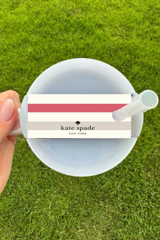 A hand holds a white cup with a white straw on green grass, featuring the Kate Spade Stripe Stanley Lid Topper by Kreative Kreationz—a chic accessory with pink, red, and beige stripes atop the lid.