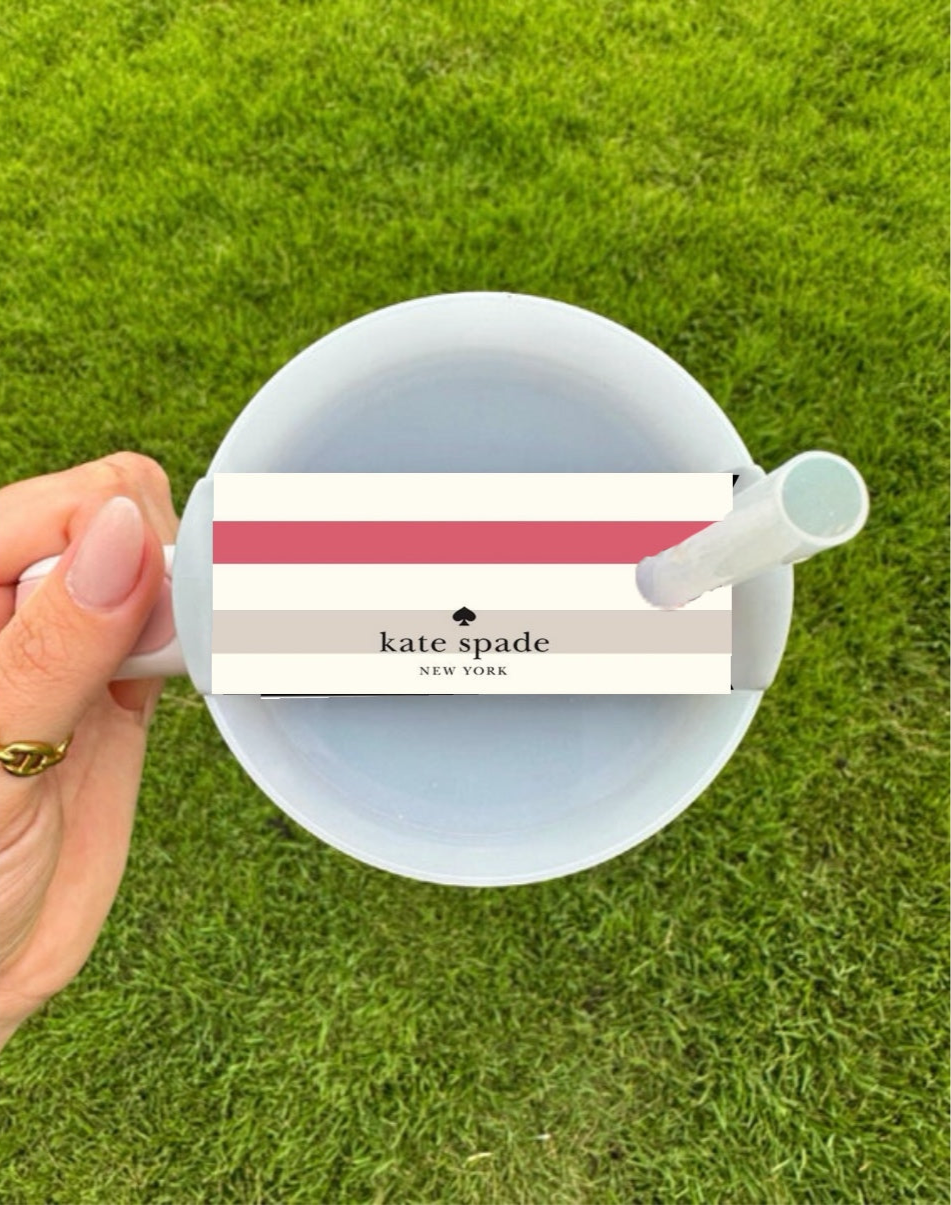 A hand holds a white cup with a white straw on green grass, featuring the Kate Spade Stripe Stanley Lid Topper by Kreative Kreationz—a chic accessory with pink, red, and beige stripes atop the lid.