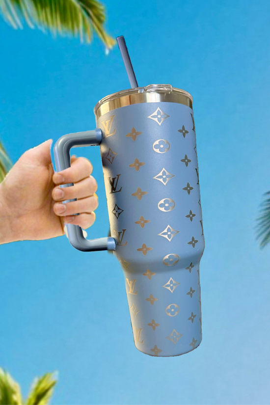 A hand holds the Kreative Kreationz LV Inspired 40oz Grey-Blue Engraved Tumbler, featuring a Louis Vuitton-inspired pattern, against a bright blue sky and palm tree branches.
