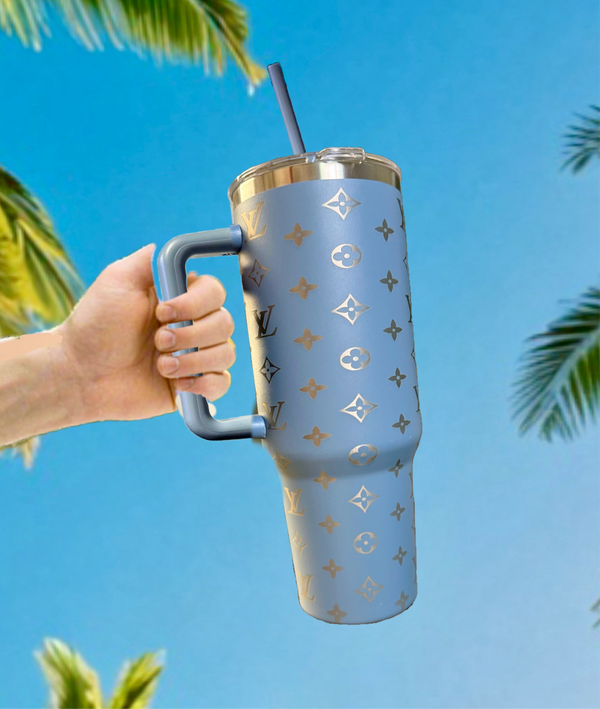 A hand holds the Kreative Kreationz LV Inspired 40oz Grey-Blue Engraved Tumbler, featuring a Louis Vuitton-inspired pattern, against a bright blue sky and palm tree branches.