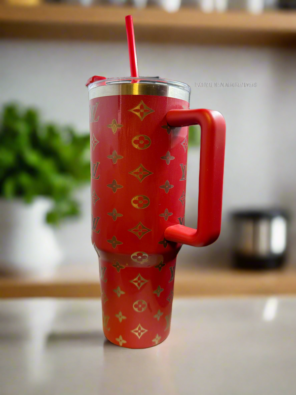 A Kreative Kreationz Louis Vuitton Inspired Gold Etched Engraved 40oz Stainless Steel Tumbler with handle and red straw sits on a countertop, with blurred kitchen items in the background.