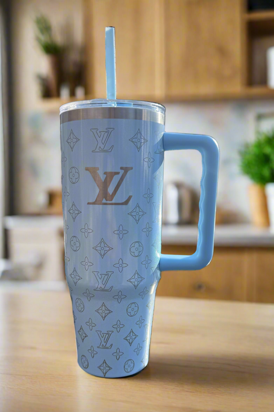 The LV Sky Blue Engraved Tumbler by Kreative Kreationz is a tall, light blue custom laser-engraved cup with a handle and straw, featuring Louis Vuitton-inspired patterns, shown on a wooden kitchen counter against a blurred background.