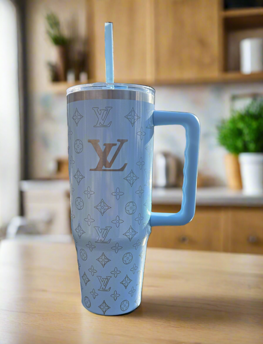 The LV Sky Blue Engraved Tumbler by Kreative Kreationz is a tall, light blue custom laser-engraved cup with a handle and straw, featuring Louis Vuitton-inspired patterns, shown on a wooden kitchen counter against a blurred background.