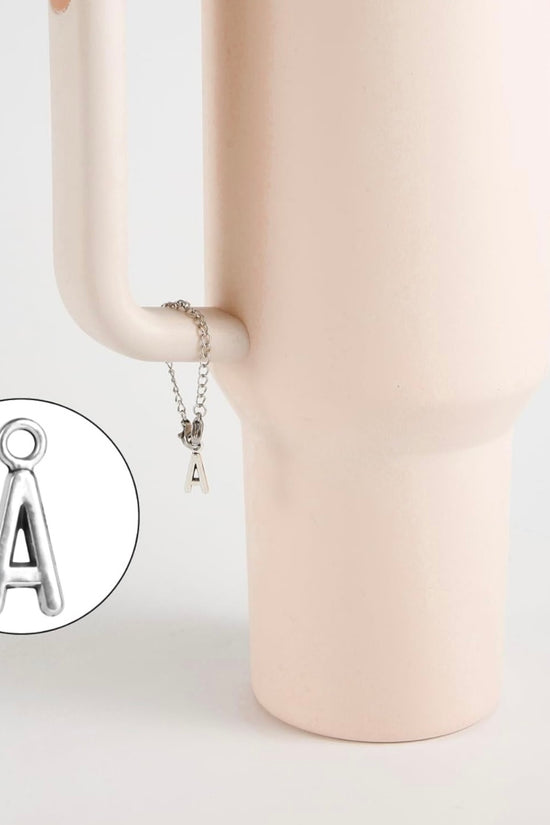 A silver Letter Cup Charm Dangle by Kreative Kreationz hangs from the handle of a matte light pink vase. An inset highlights the 