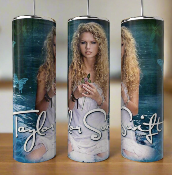 The Taylor Swift Debut 20oz Tumbler by Kreative Kreationz features her with curly hair in a white dress, holding a flower against blue water with butterflies and her signature. Stainless steel, eco-friendly & reusable.