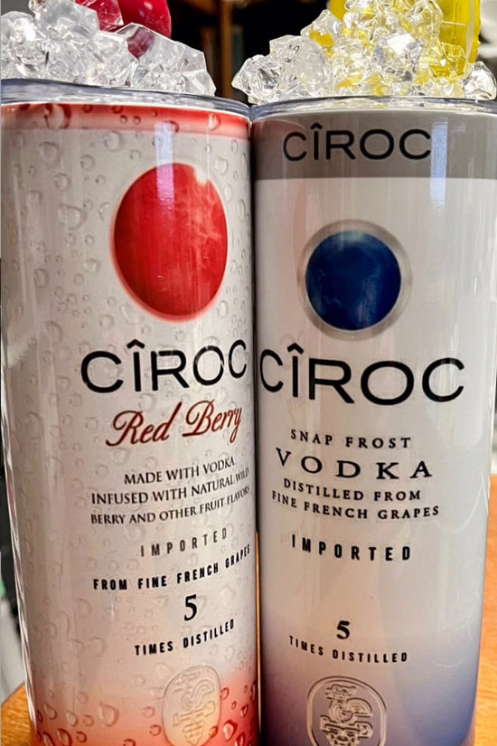A lineup of CÎROC vodka cans in various flavors including original, mango, red berry, coconut, watermelon, and peach is beautifully arranged on an outdoor surface with a green hedge and blue sky background. Each can is paired with a chic 20oz Ciroc Tumbler with 3D Lid Topper by Kreative Kreationz.