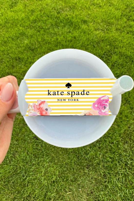 A hand holds a white tea cup and saucer beside the Kate Spade Spring Stripe Stanley Lid Topper by Kreative Kreationz, featuring yellow and white stripes with floral accents, set against green grass. Perfect for pairing with your favorite Stanley accessory.