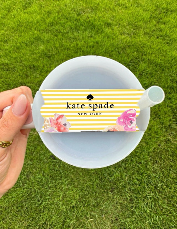 A hand holds a white tea cup and saucer beside the Kate Spade Spring Stripe Stanley Lid Topper by Kreative Kreationz, featuring yellow and white stripes with floral accents, set against green grass. Perfect for pairing with your favorite Stanley accessory.