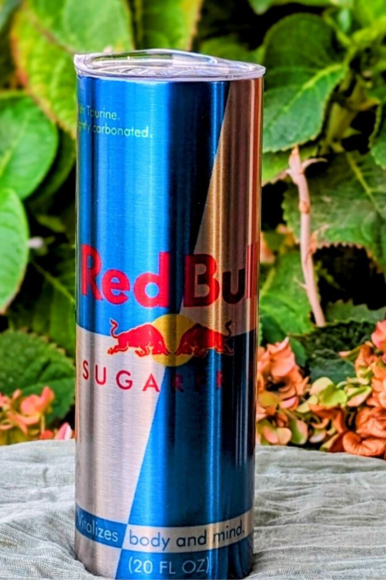 A 20 oz RB Energy-Silver Stainless Steel Tumbler from Kreative Kreationz, resembling a Red Bull Sugar Free can, stands on fabric with leafy greens and pink flowers in the background. The silver and blue tumbler features the Red Bull design, highlighting its revitalizing benefits.