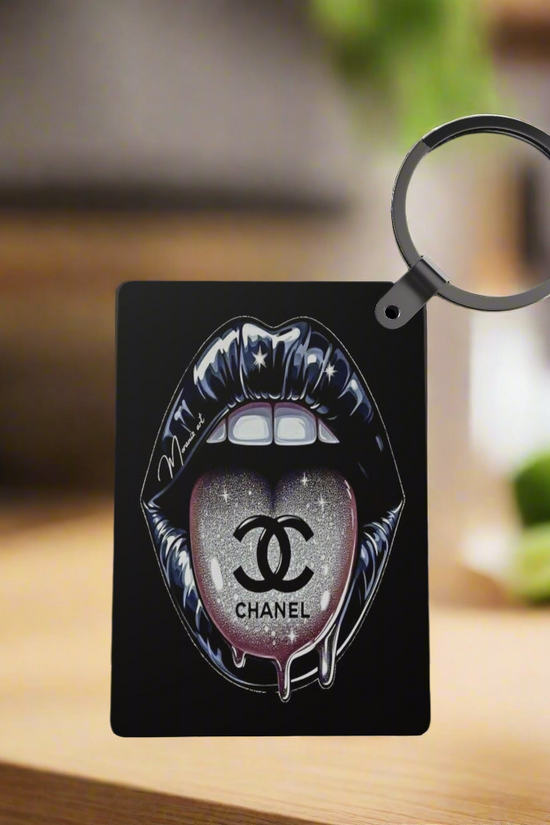 A black card displays glossy, stylized Chanel lips with a glittery logo on the tongue. Attached is the Chanel Inspired Keychain by Kreative Kreationz—a round black sublimation keychain—set against a softly blurred background.