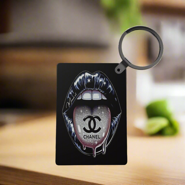 A black card displays glossy, stylized Chanel lips with a glittery logo on the tongue. Attached is the Chanel Inspired Keychain by Kreative Kreationz—a round black sublimation keychain—set against a softly blurred background.