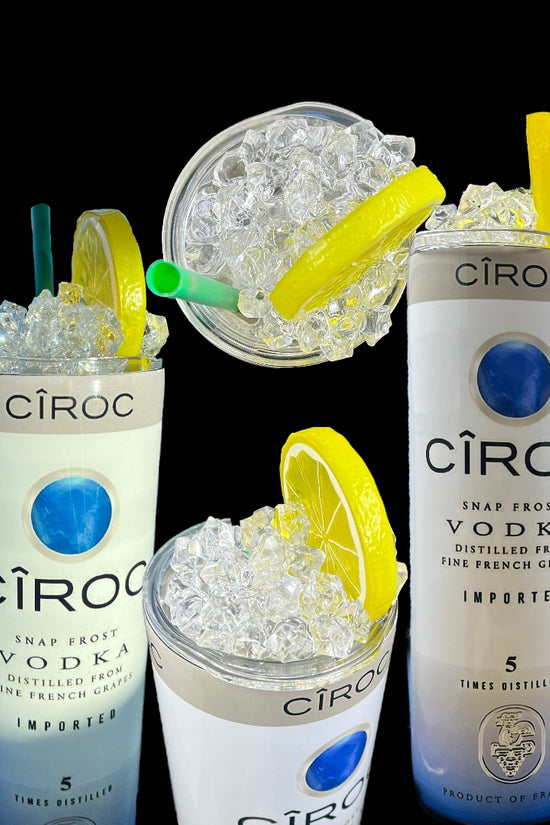 Two cans of Kreative Kreationz Ciroc 20oz Tumbler with 3D Lid Topper are shown on a wooden surface, reminiscent of travel tumblers. The left can is Red Berry flavor, topped with ice and a cherry. The right can is Vodka Snap Frost flavor, topped with ice and a lemon slice. Both cans feature product information and branding.
