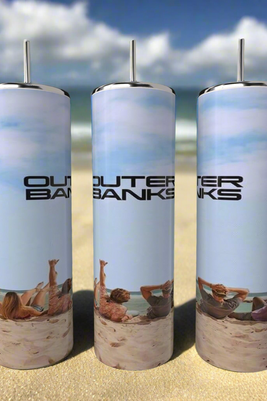 Three Outer Banks OBX 20oz Tumblers by Kreative Kreationz sit on a sandy beach, capturing the scene of beachgoers under a clear sky with ocean waves. Made of stainless steel, each tumbler is ideal for relaxing by the ocean.