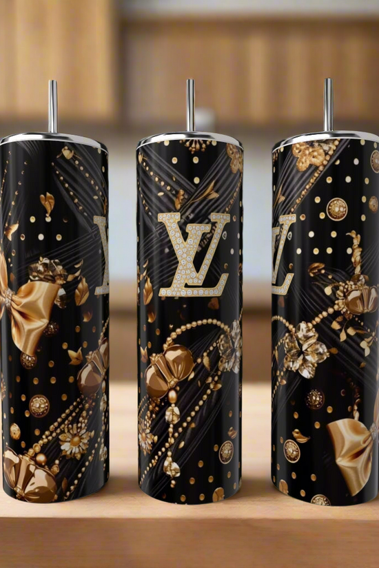 Three Louis Vuitton Black & Gold 20oz tumblers by Kreative Kreationz, featuring metallic gold and silver floral designs, bows, and the iconic LV logo, are displayed on a wooden surface in a modern kitchen.