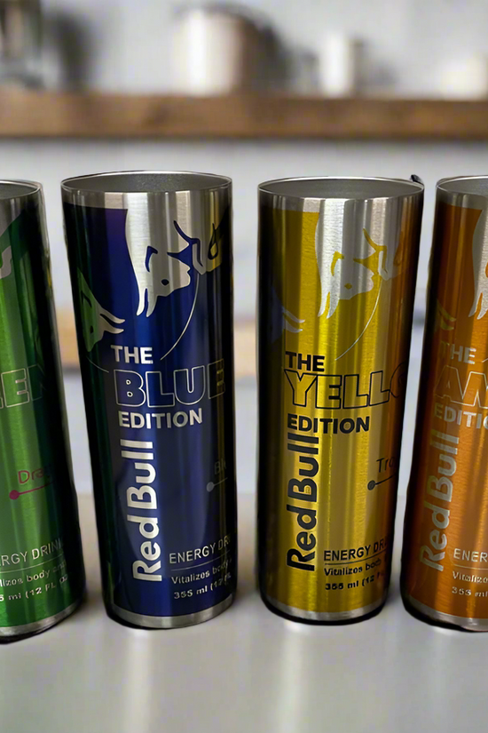 Four colorful cans of Red Bull energy drinks are lined up on a counter. From left to right, the cans are green (Green Edition, Dragon Fruit flavor), blue (Blue Edition, Blueberry flavor), yellow (Yellow Edition, Tropical flavor), and orange (Amber Edition, Strawberry-Apricot flavor). Next to them sits a stylish Red Bull Stainless Steel 20oz Tumbler by Kreative Kreationz in a chic kitchen setting.