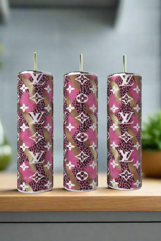 A Louis Vuitton Pink & Gold 20oz Tumbler by Kreative Kreationz featuring luxury-inspired design and repeating “LV” logo sits on a wooden surface, with pink, brown, and white patterns; a blurred kitchen appears in the background.