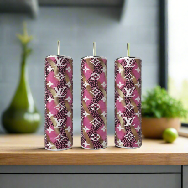 A Louis Vuitton Pink & Gold 20oz Tumbler by Kreative Kreationz featuring luxury-inspired design and repeating “LV” logo sits on a wooden surface, with pink, brown, and white patterns; a blurred kitchen appears in the background.
