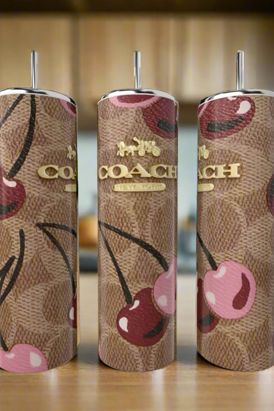 Three Cherry Brown Coach 20oz Skinny Tumblers by Kreative Kreationz, featuring a stainless steel build, brown monogram, pink cherry designs, silver straws, and gold logos, are displayed on a wooden surface with a blurred kitchen background.