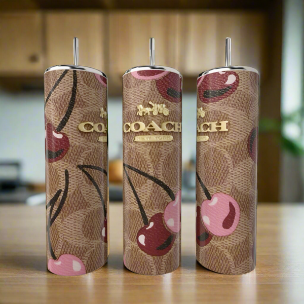 Three Cherry Brown Coach 20oz Skinny Tumblers by Kreative Kreationz, featuring a stainless steel build, brown monogram, pink cherry designs, silver straws, and gold logos, are displayed on a wooden surface with a blurred kitchen background.