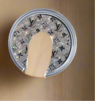 The LV Shimmer Skinny Lid Topper by Kreative Kreationz features a glittery Louis Vuitton-inspired pattern and comes partially opened with a flat wooden spoon, ideal as a sublimatable aluminum topper for skinny tumblers on a plain brown surface.