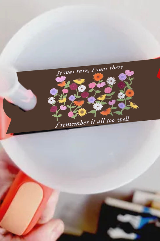 A close-up image features a Kreative Kreationz Taylor Swift Debut Piano Stanley Lid Plate/Topper: round, white with an orange handle, adorned with flowers and a brown banner reading 