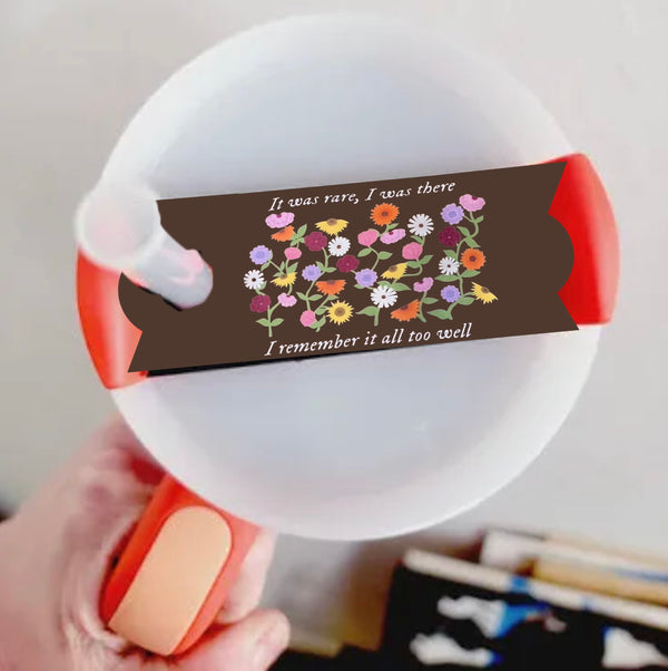 A close-up image features a Kreative Kreationz Taylor Swift Debut Piano Stanley Lid Plate/Topper: round, white with an orange handle, adorned with flowers and a brown banner reading "It was rare, I was there I remember it all too well," held by a hand against a blurred background.