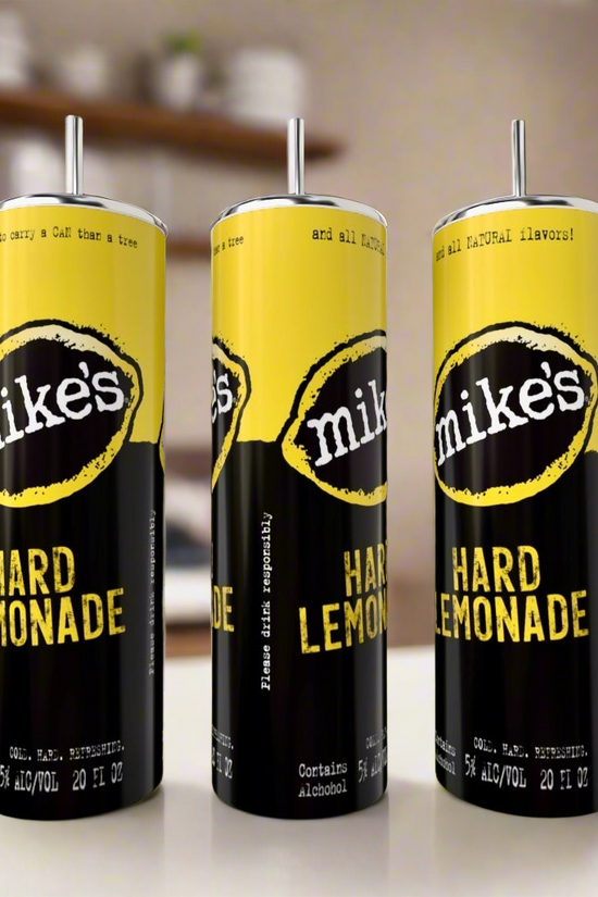 Three Mikes Hard Lemonade cans with yellow and black labels are displayed beside a Kreative Kreationz Mikes Hard Lemonade Variety 20oz eco-friendly stainless steel tumbler, set against a blurred kitchen backdrop with shelves and dishes.