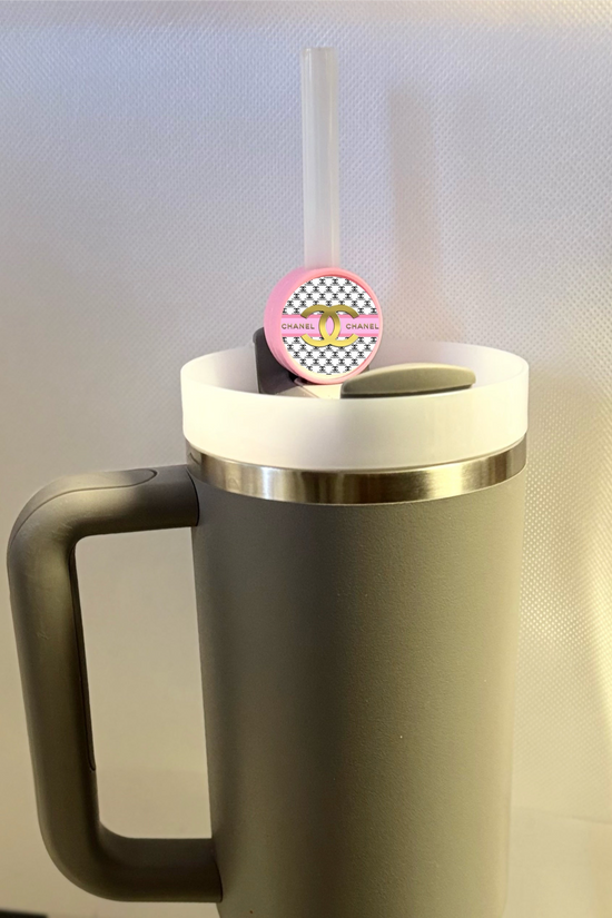 A gray mug with a handle, featuring a white lid with a straw, is accessorized by a Kreative Kreationz Chanel Straw Topper. The silicone round topper showcases a pink fishnet pattern and the word 