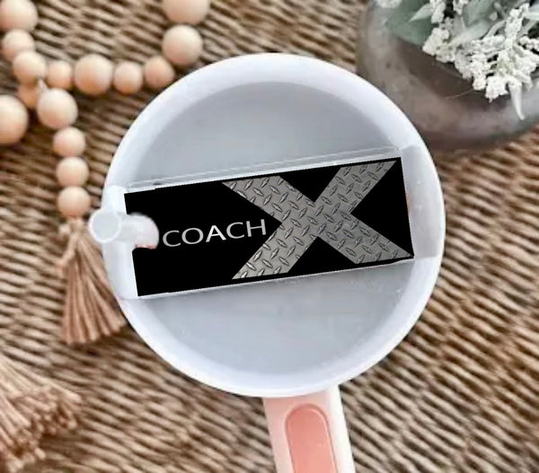 A Kreative Kreationz X Coach | Stanley Lid Plate/Topper decorates a white cup with liquid, featuring a "COACH X" label and metallic X design. The cup sits on a woven surface near wooden beads and a plant.