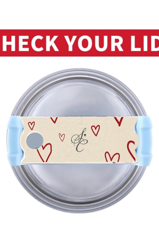 A silver tumbler filled with black liquid features a beige piece of tape adorned with red hearts and a cursive letter 