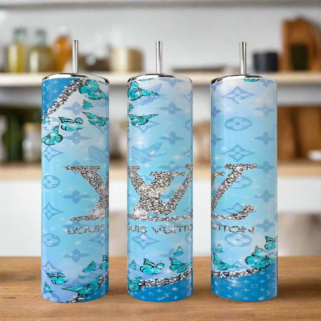 Three Kreative Kreationz Butterfly LV 20oz Tumblers with metal straws are shown on wood. Each features a luxury-inspired blue gradient with glittery "LV," butterflies, and elegant motifs, set against a blurred kitchen background.