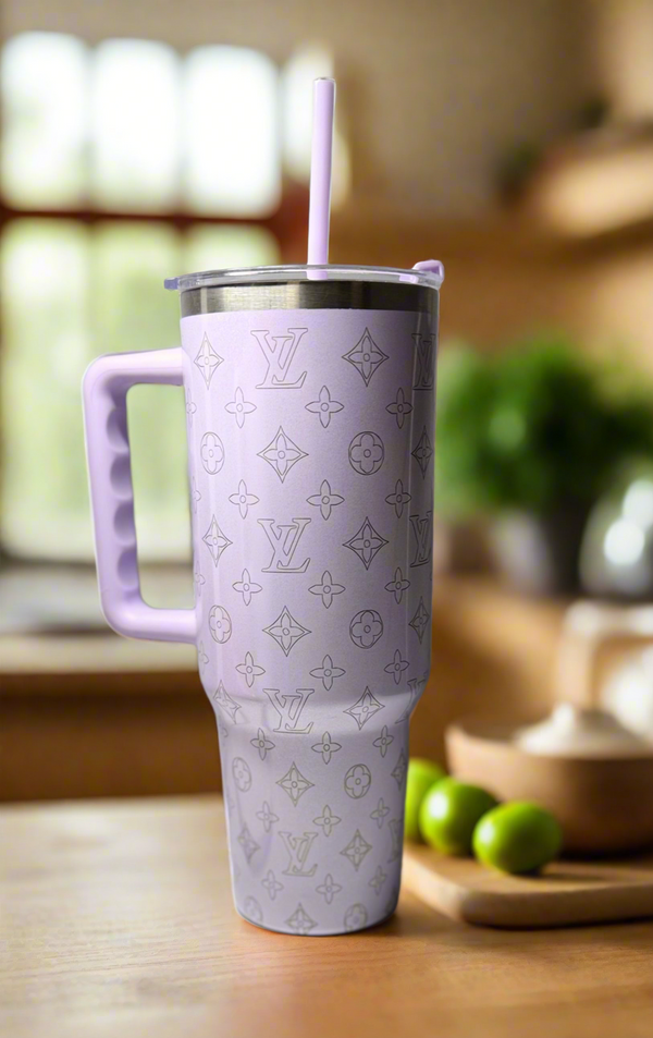 The LV Lilac Engraved Tumbler by Kreative Kreationz, featuring a handle, straw, and Louis Vuitton-inspired monogram design, sits on a wooden kitchen counter with limes and kitchen items blurred in the background.