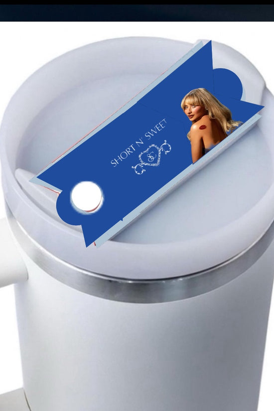 A white mug with a light blue button feature, covered by a lid. On the lid sits a blue, rectangular packaging featuring an image of a woman with blonde hair in a black outfit. The text on the packaging reads 