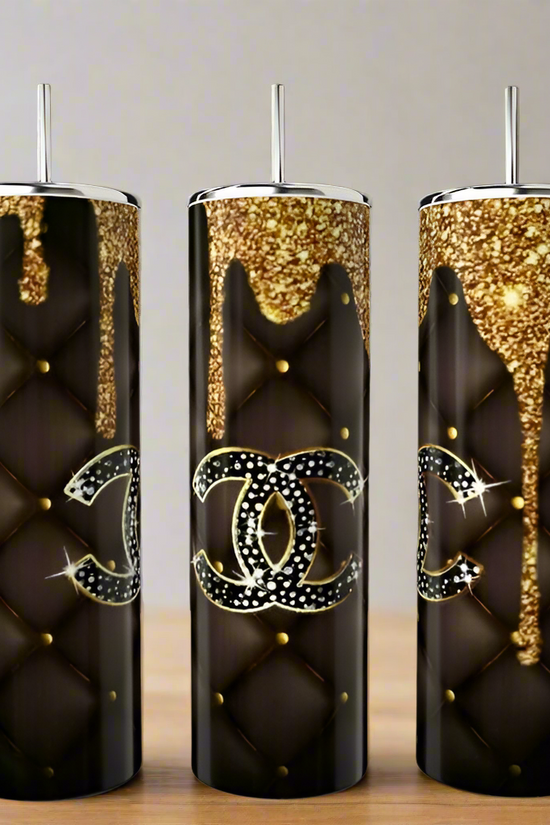 Three tall, black Chanel Gold Drip 20oz Tumblers by Kreative Kreationz feature quilted patterns, gold studs, glitter accents at the top, and a rhinestone double C logo, displayed on a wooden surface.