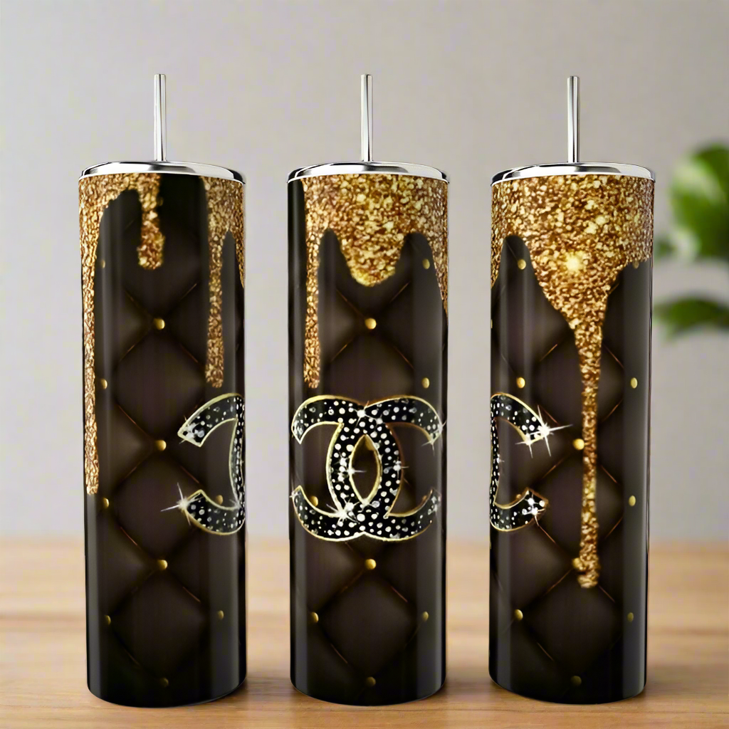 Three tall, black Chanel Gold Drip 20oz Tumblers by Kreative Kreationz feature quilted patterns, gold studs, glitter accents at the top, and a rhinestone double C logo, displayed on a wooden surface.