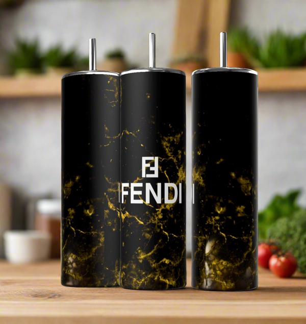 Three FENDI Black Gold 20oz Skinny Tumblers by Kreative Kreationz, featuring a black and gold marble design with the iconic "Fendi" logo, sit on a wooden table. In this warm kitchen setting with blurred vegetables, their double-wall insulation keeps drinks at the perfect temperature.