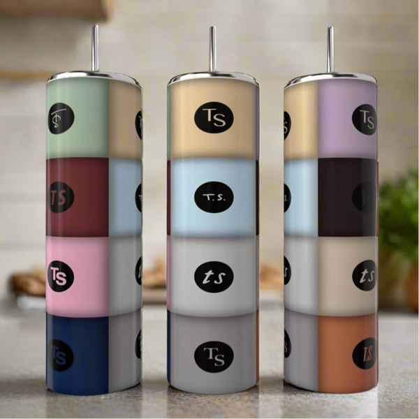 Three Kreative Kreationz TA12 Taylor Swift 20oz stainless steel tumblers with metal lids and straws, each featuring colorful "Ts" or "T.S." segments, are stacked on a kitchen countertop with a blurred background.