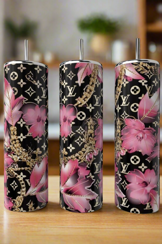 Three LV Pink 20oz Tumblers by Kreative Kreationz, crafted from stainless steel, feature metal straws and stand on a wooden counter. With a black background adorned with pink floral and gold chain designs plus monogram patterns, these eco-friendly beauties enhance any blurred kitchen setting.