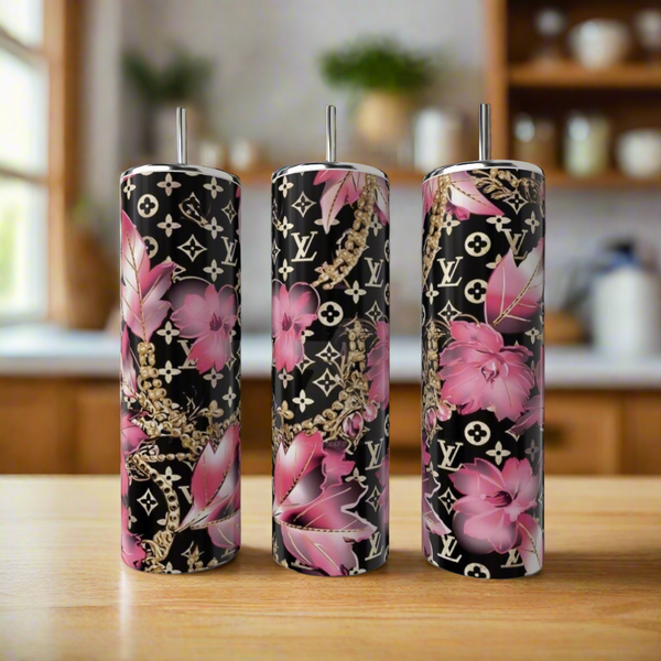 Three LV Pink 20oz Tumblers by Kreative Kreationz, crafted from stainless steel, feature metal straws and stand on a wooden counter. With a black background adorned with pink floral and gold chain designs plus monogram patterns, these eco-friendly beauties enhance any blurred kitchen setting.