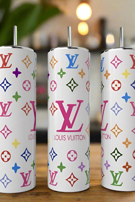 Three sleek, white Louis V Multicolor tumblers by Kreative Kreationz, each 20oz with colorful logos, rest on a wooden table. The stainless steel finish offers a luxury-inspired design against a blurred background that suggests a cozy setting with green foliage.
