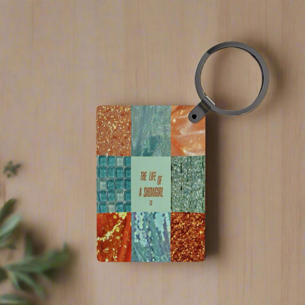 A designer-inspired "The Life of a Showgirl Inspired Keychain" by Kreative Kreationz rests on a wooden surface beside a colorful book, a magnifying glass, and green plant leaves in the corner.