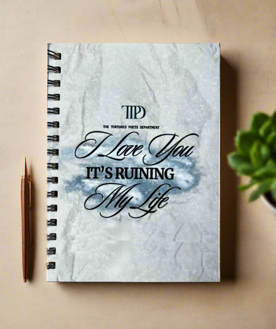 The Kreative Kreationz TTPD A6 Notebook features a marble-textured cover with "I Love You IT'S RUINING My Life" in elegant script and a sturdy spiral binding, shown beside a gold pen and green potted plant on a beige surface.