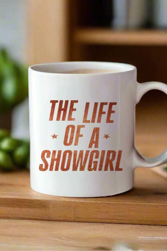 The Life Of a Showgirl Mug by Kreative Kreationz, featuring bold orange lettering, sits on a wooden surface with blurred green vegetables and kitchen items in the background.