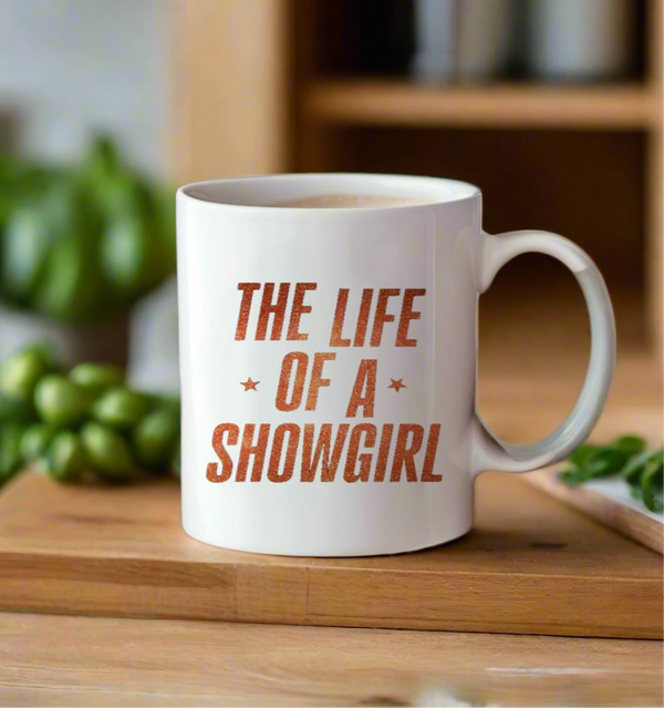 The Life Of a Showgirl Mug by Kreative Kreationz, featuring bold orange lettering, sits on a wooden surface with blurred green vegetables and kitchen items in the background.