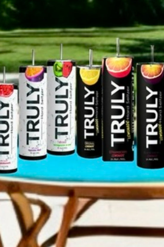 Cans of Truly hard seltzer in an array of flavors sit beside a Kreative Kreationz Truly Tumbler, featuring a 3D fruit and ice lid topper, with a scenic pool and lush greenery in the background.