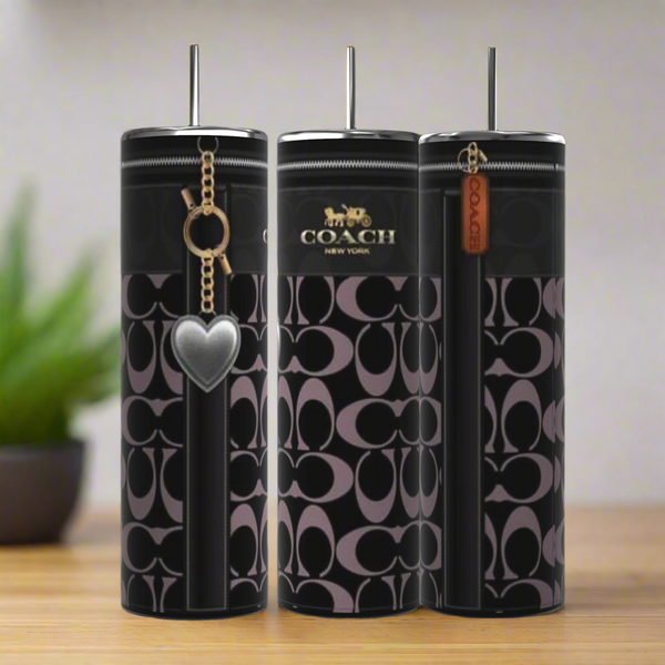 Three black Kreative Kreationz Coach Heart Purse 20oz Tumblers with Coach logo and monogram pattern sit on wood. Two have keychain accessories—a heart charm and leather tag. A blurred green plant appears in the background.