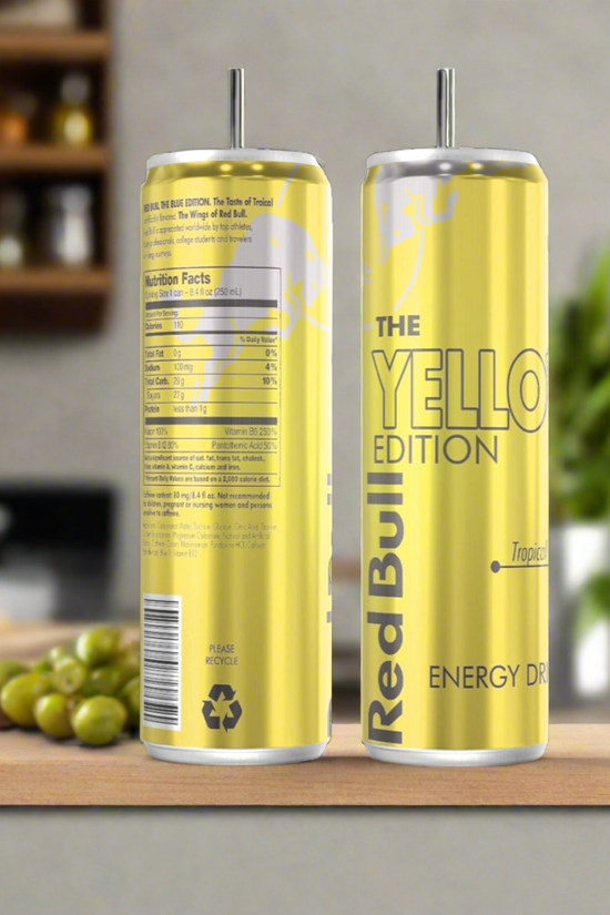 A Yellow Tropical Red Bull 20oz Tumbler by Kreative Kreationz sits on a wooden surface beside two yellow Red Bull cans—one labeled 